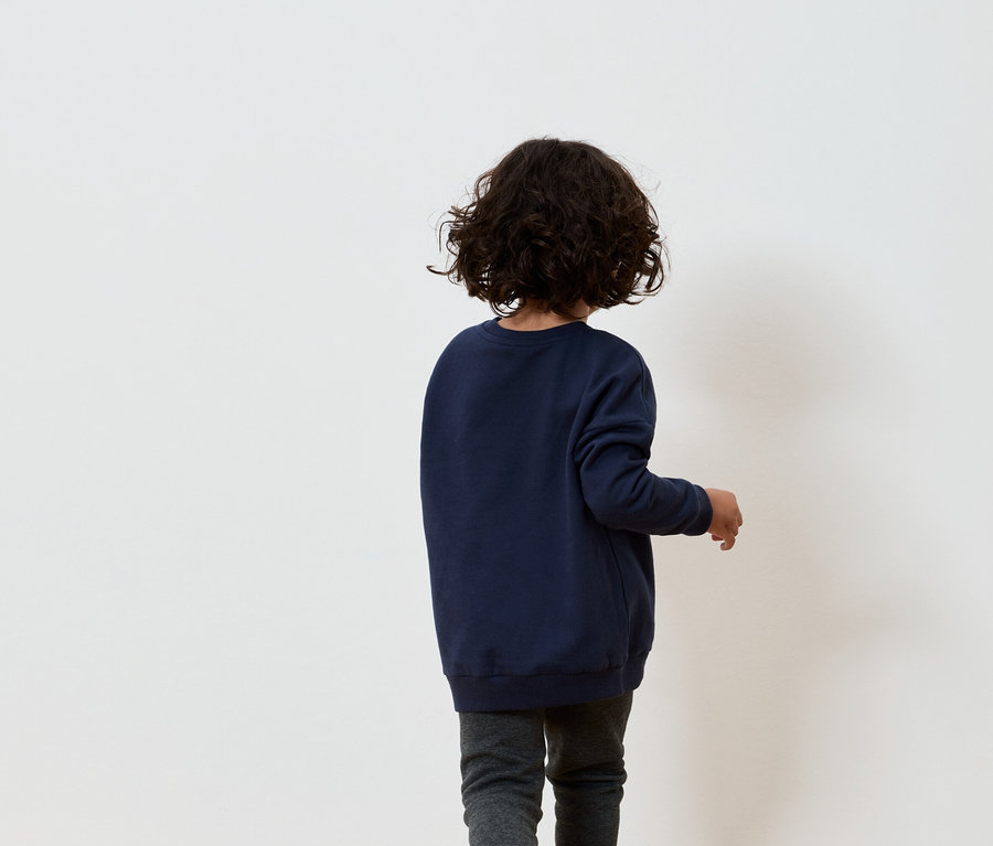 Chlapec s hnedými kučeravými vlasmi v tmavomodrej mikine a sivých nohaviciach stojí chrbtom k bielej stene.