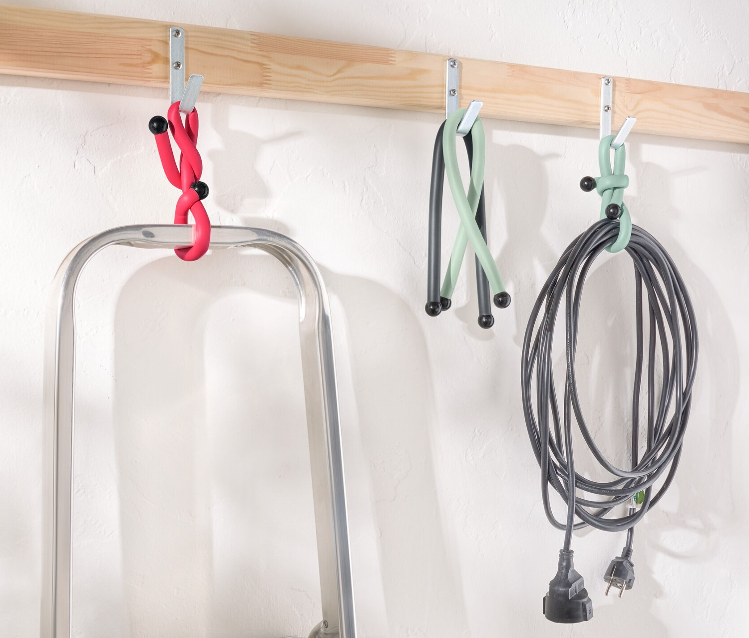Rebrík stojí pod drevenou lištou s háčikmi, na ktorých visia Power Kabelbinder v červenej, zelenej a čiernej farbe.