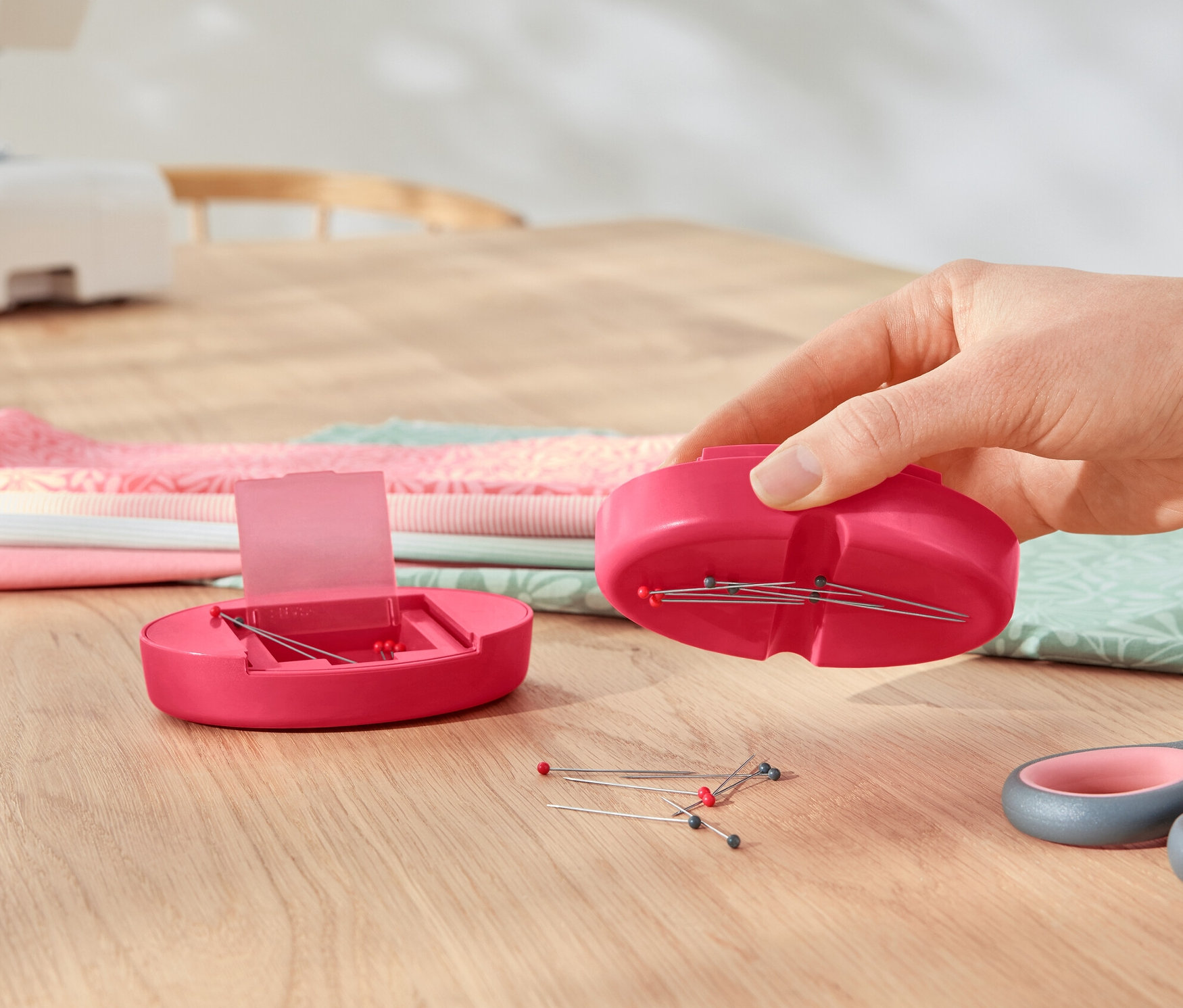 Ruka drží magnetický držiak ihiel nad otvoreným magnetickým držiakom ihiel, vedľa ležia nožnice a hromada látok.