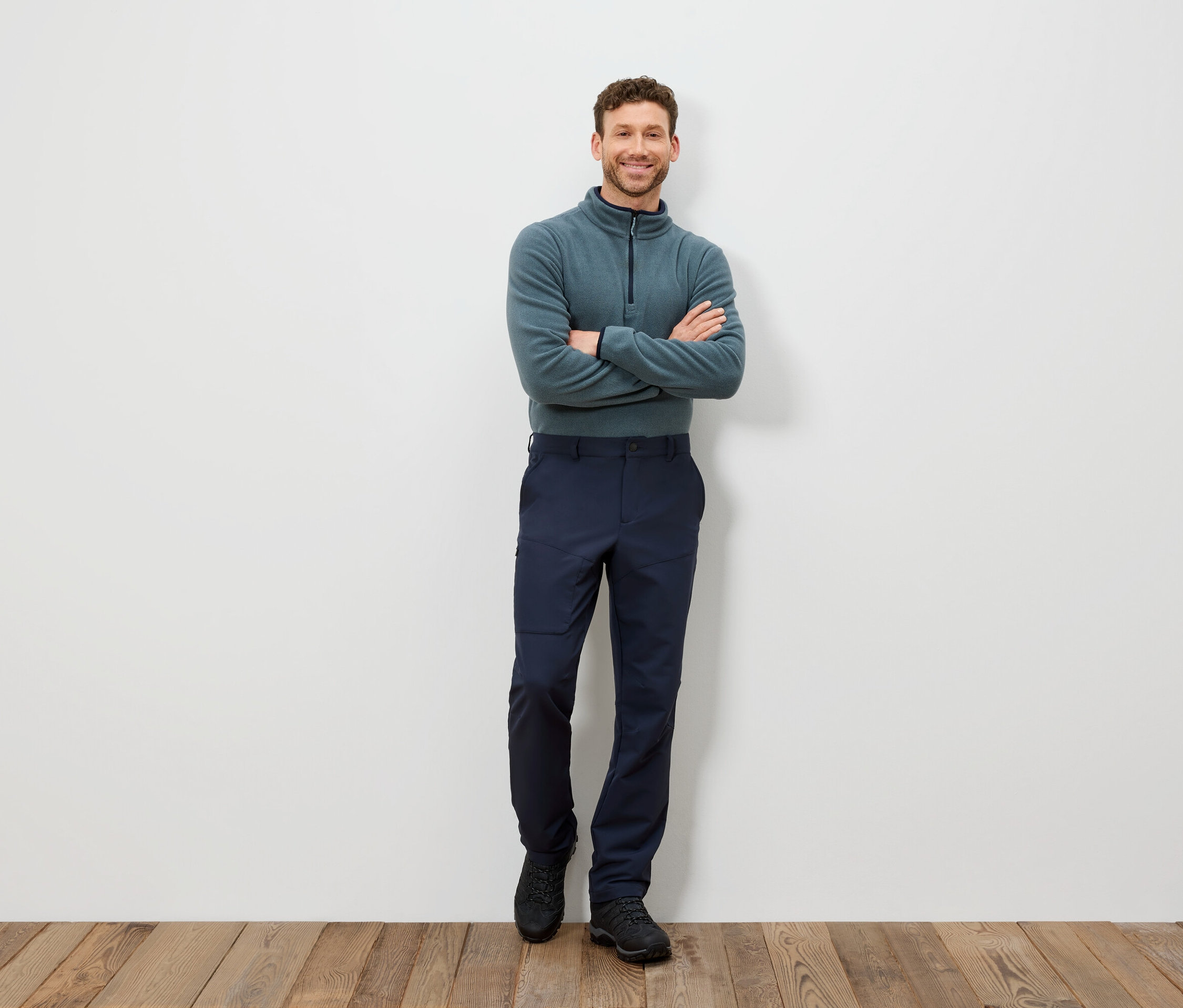 Muž stojí s prekríženými rukami na hrudi, má na sebe modrozelený pulóver a tmavomodré nohavice.