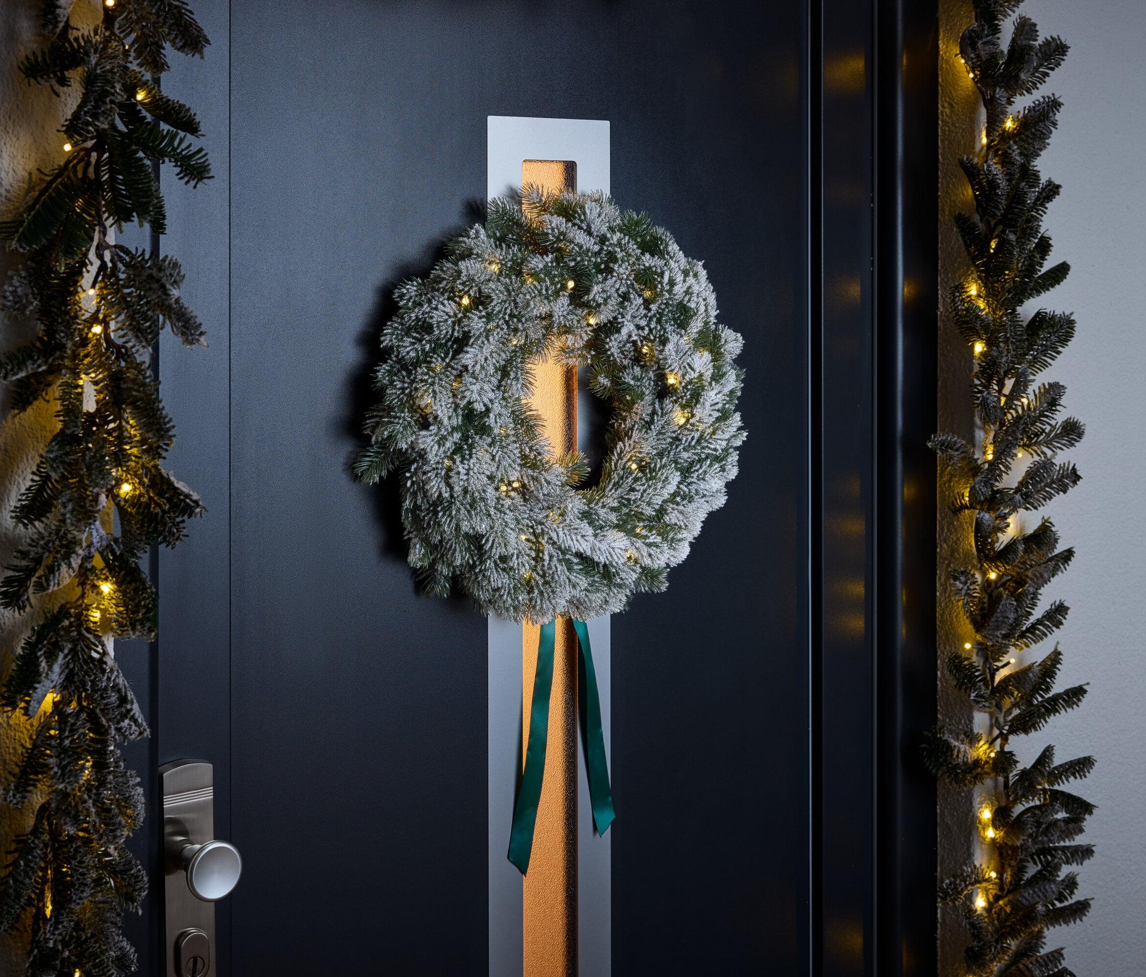 Venček s bielym posypom a zelenou stuhou visí na tmavomodrých dverách. Girlandy s malými svetielkami lemujú dvere.
