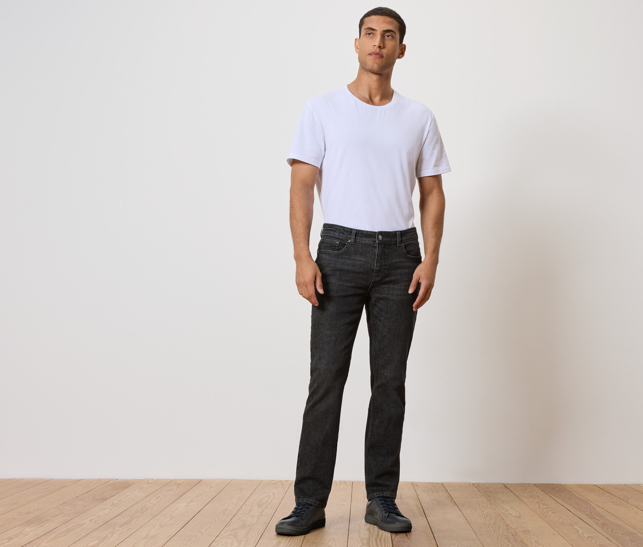 Muž stojí na drevenej podlahe a má na sebe čierne džínsy a biele tričko.
