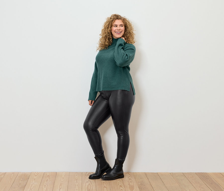 Žena s kučeravými vlasmi pózuje v zelenom roláku, čiernych koženkových legínach a čiernych topánkach.