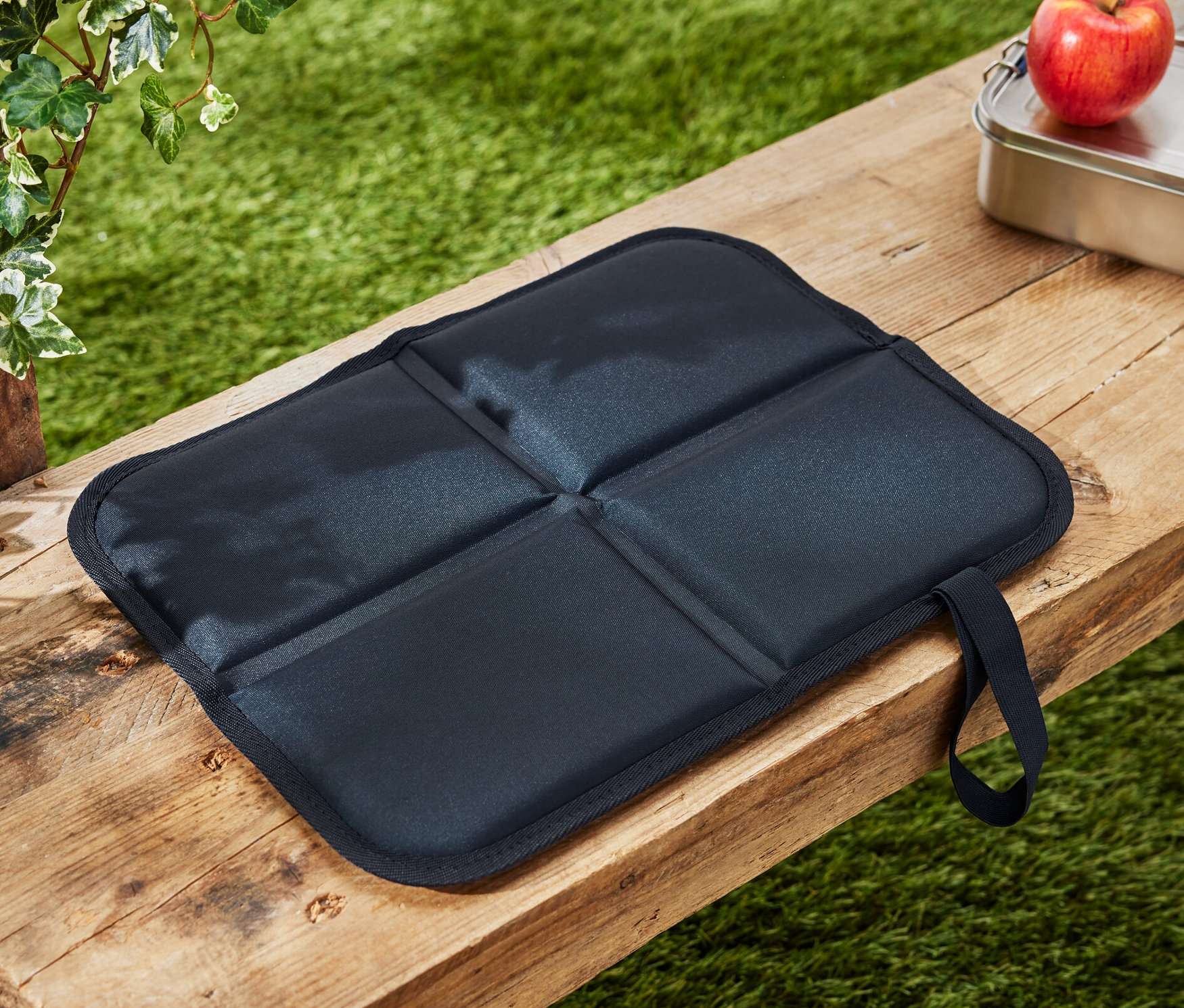 Skladací termovankúš na sedenie leží na drevenej lavici vedľa jablka a desiatového boxu.