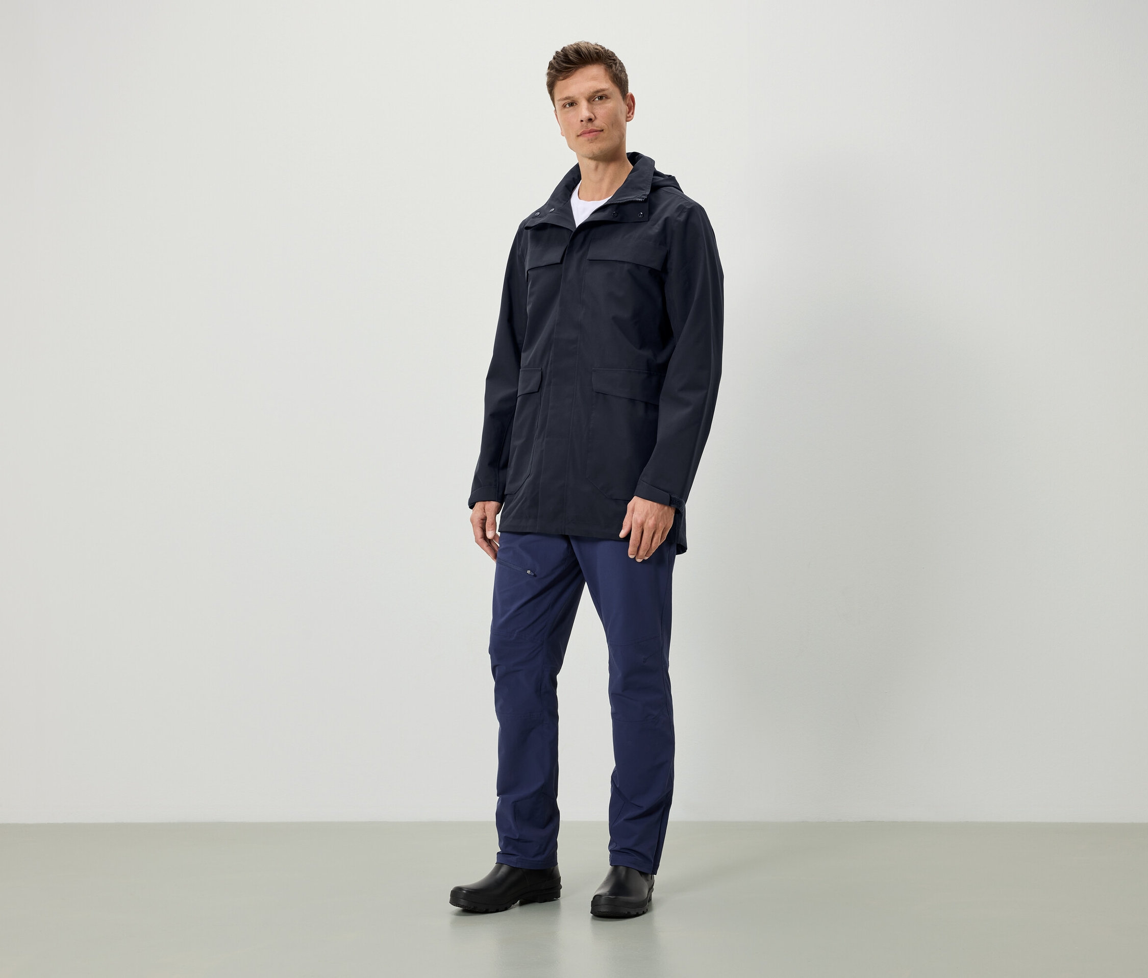 Muž stojí a má na sebe tmavomodrú bundu s kapucňou, tmavomodré nohavice a čierne topánky.