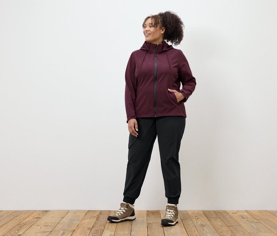 Žena v bordovej bunde na zips, čiernych nohaviciach a hnedých topánkach stojí na drevenej podlahe.