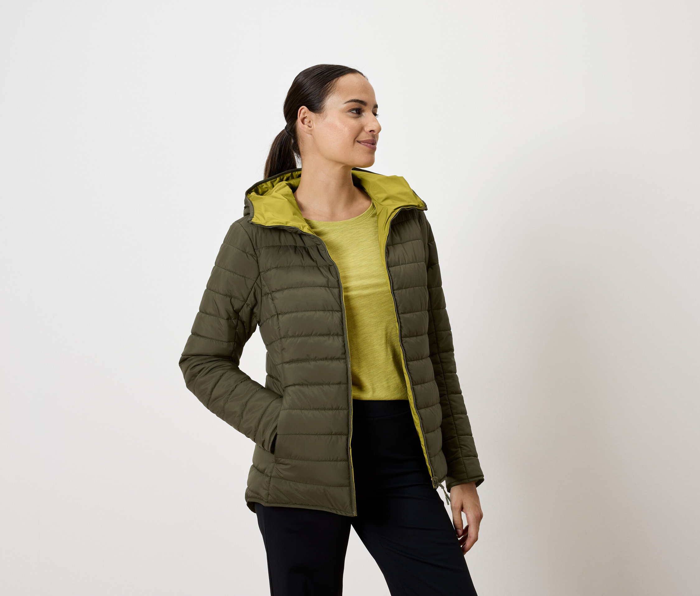 Žena v olivovozelenej prešívanej bunde s kapucňou, ruky vo vreckách, pozerá sa doprava.