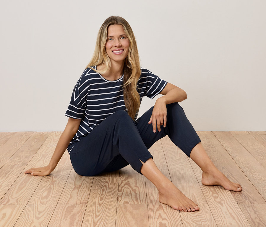 Blondína sedí na drevenej podlahe v námorníckom pruhovanom tričku a tmavomodrých nohaviciach.