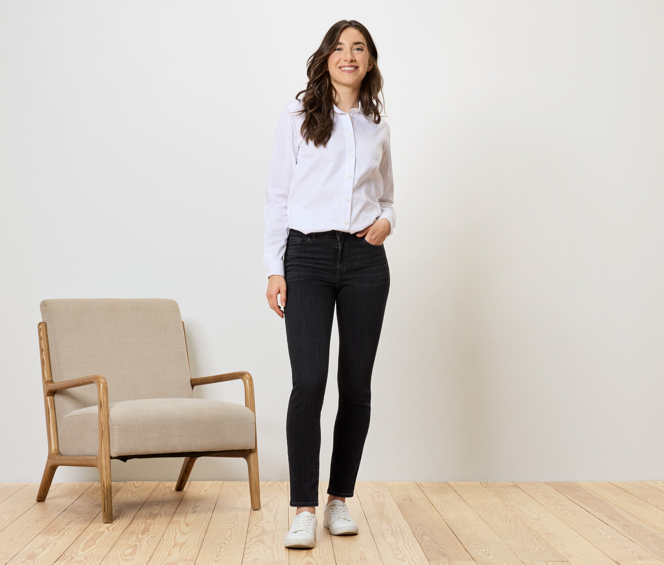 Žena stojí v čiernych džínsoch, bielej košeli a bielych topánkach vedľa béžového kresla.