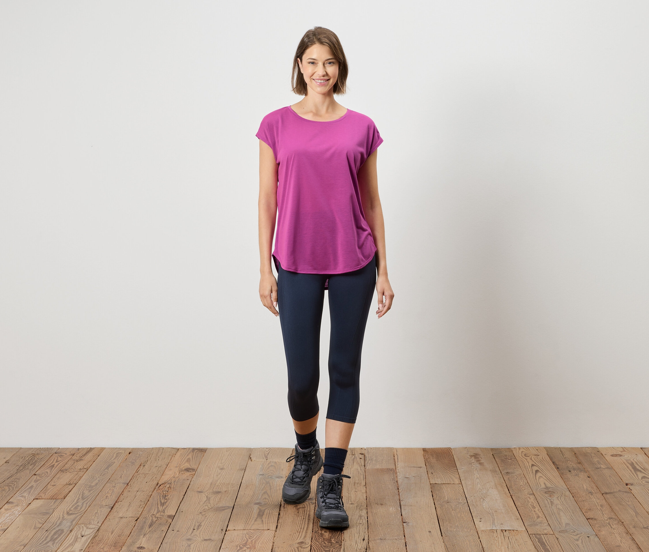 Žena stojí na drevenej podlahe pred bielou stenou. Má na sebe ružové tričko, modré legíny a sivé topánky.