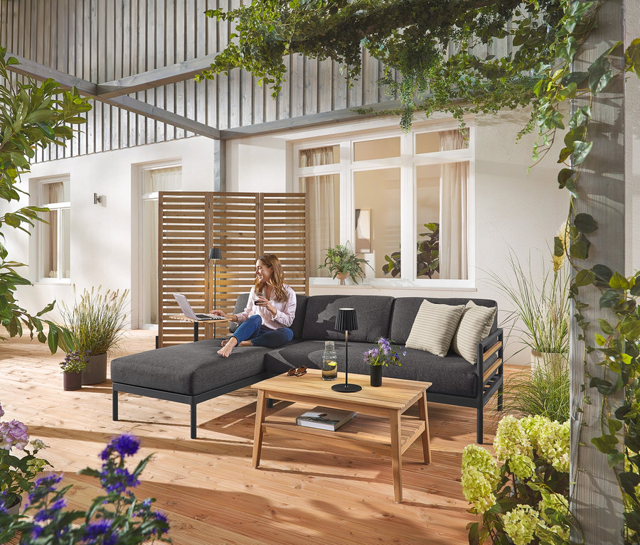 Žena sedí na záhradnej rohovej súprave Premium »Elin« s flexibilným paravánom v pozadí. Vidieť aj čierne LED akumulátorové vonkajšie stojacie a stolové svietidlo a záhradný konferenčný stolík.