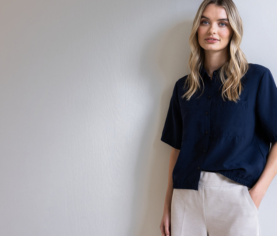 Žena s blond vlasmi v tmavomodrej košeli a béžových nohaviciach pózuje pred svetlosivou stenou.