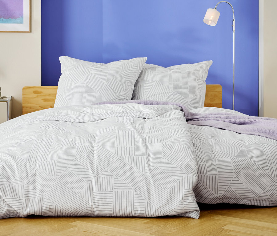 Posteľ s bielymi obliečkami s geometrickým vzorom a dvoma vankúšmi stojí pred modrou stenou.