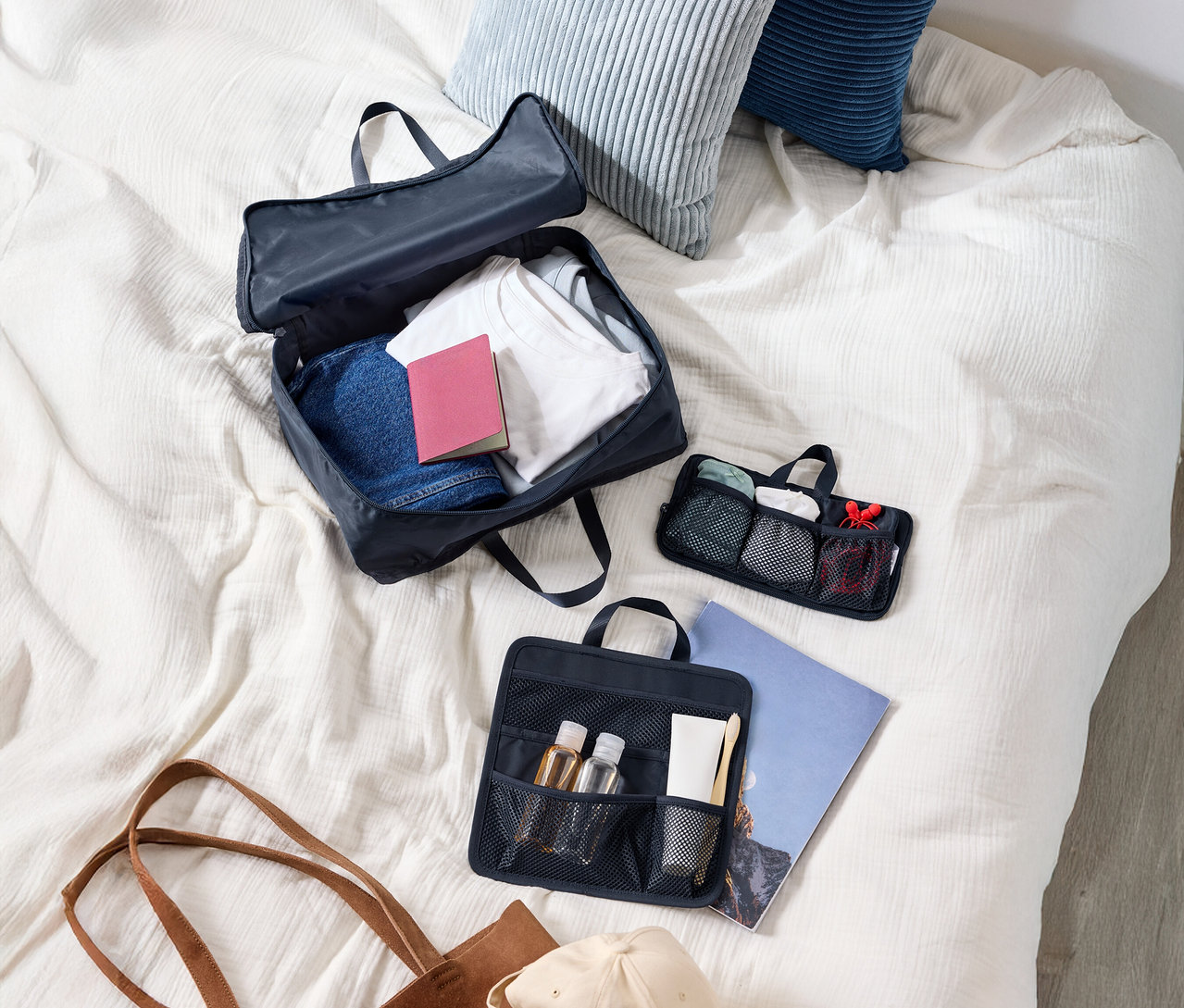 Pohľad zhora na kufor s oblečením, dva vankúše, kozmetickú tašku, tašku a čiapku na posteli.