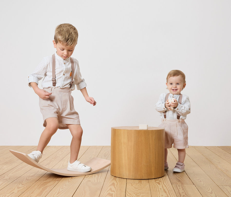 Dve deti, chlapec balansuje na drevenej doske, dojča stojí vedľa dreveného stolíka a drží hračku.