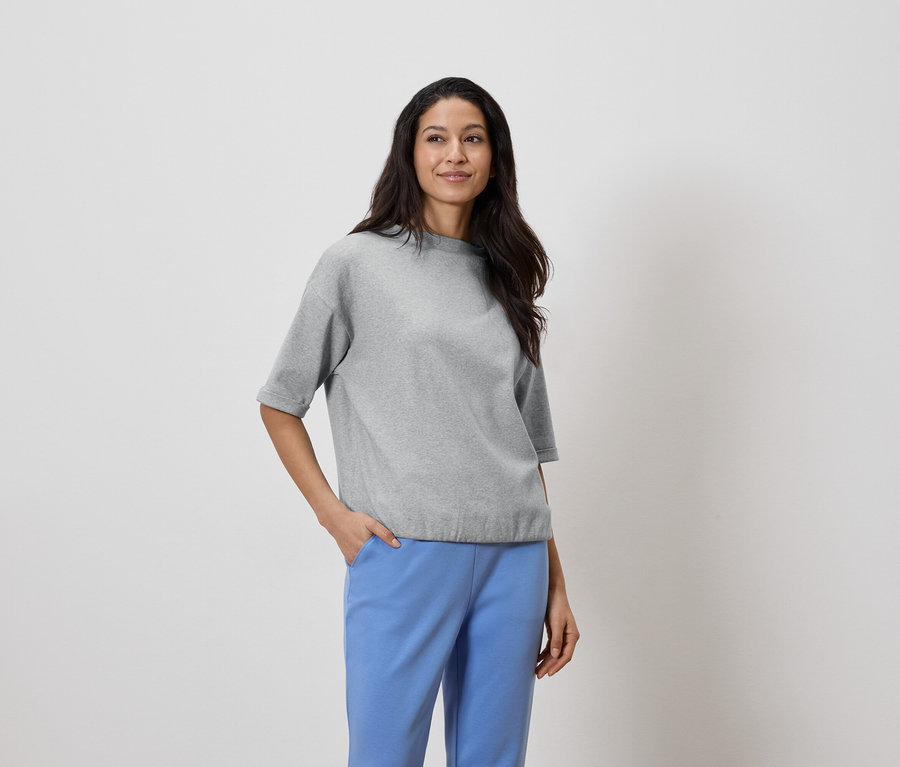 Žena s tmavými vlasmi má na sebe sivý top a modré nohavice, ruka vo vrecku.