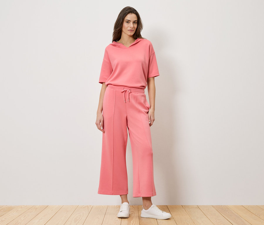 Žena stojí pred stenou a má na sebe koralový dvojdielny outfit pozostávajúci z mikiny s krátkym rukávom a culottes nohavíc spolu s bielymi teniskami.