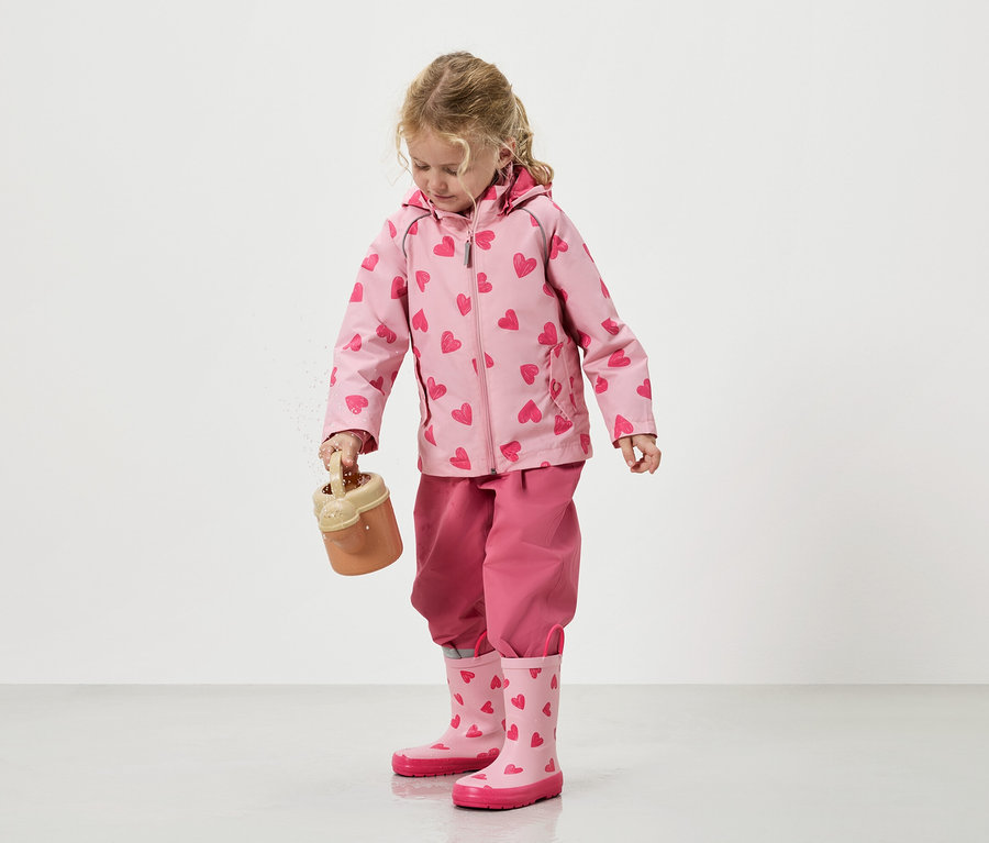 Dievčatko sype piesok z hračkárskeho vedierka, oblečené v ružovej bundičke so vzorom srdiečok, zladených nohaviciach a gumákoch.