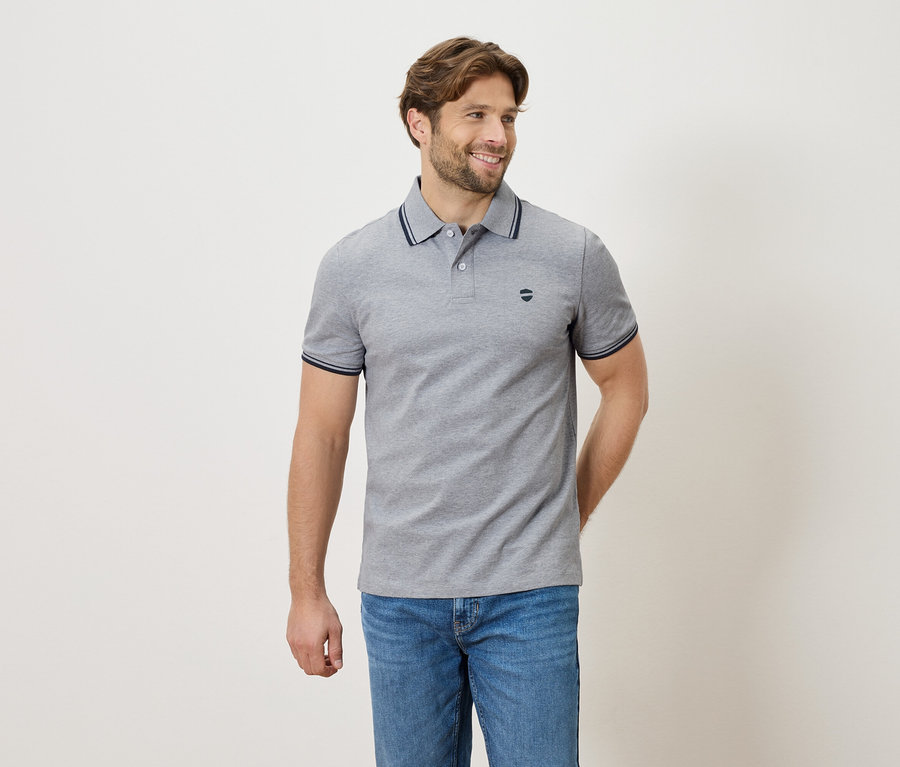 Muž v šedom polo tričku a džínsoch stojí na neutrálnom pozadí.