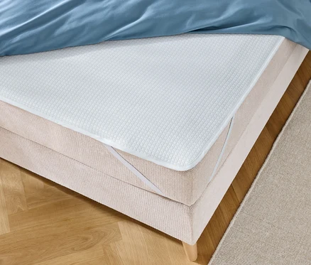 Detailný záber postele s modrou posteľnou bielizňou a bielym chráničom matraca s elastickými pásmi.