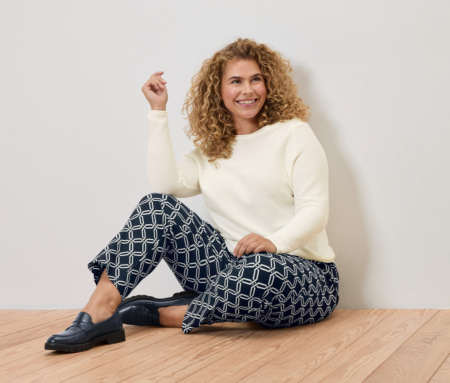 Žena s kučeravými vlasmi sedí na drevenej podlahe, má na sebe krémový sveter a modré nohavice s geometrickými vzormi.