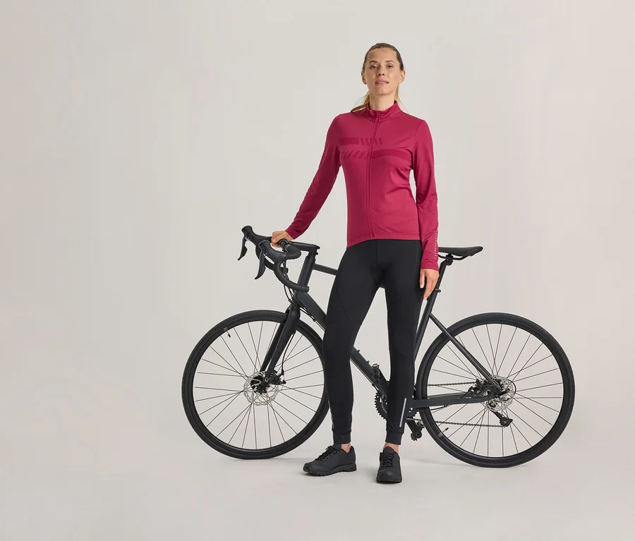 Žena stojí vedľa čierneho cestného bicykla, má na sebe ružový cyklistický dres s dlhým rukávom, čierne cyklistické nohavice a čierne topánky.