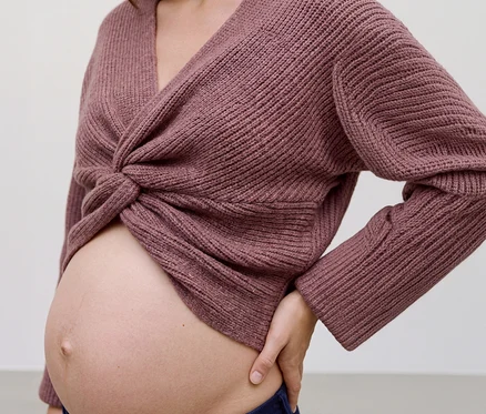 Tehotná žena má na sebe ružový pletený top MAMALICIOUS »SVALA« s prekríženou prednou časťou, ktorý odhaľuje jej brucho. Jej ľavá ruka spočíva na boku.