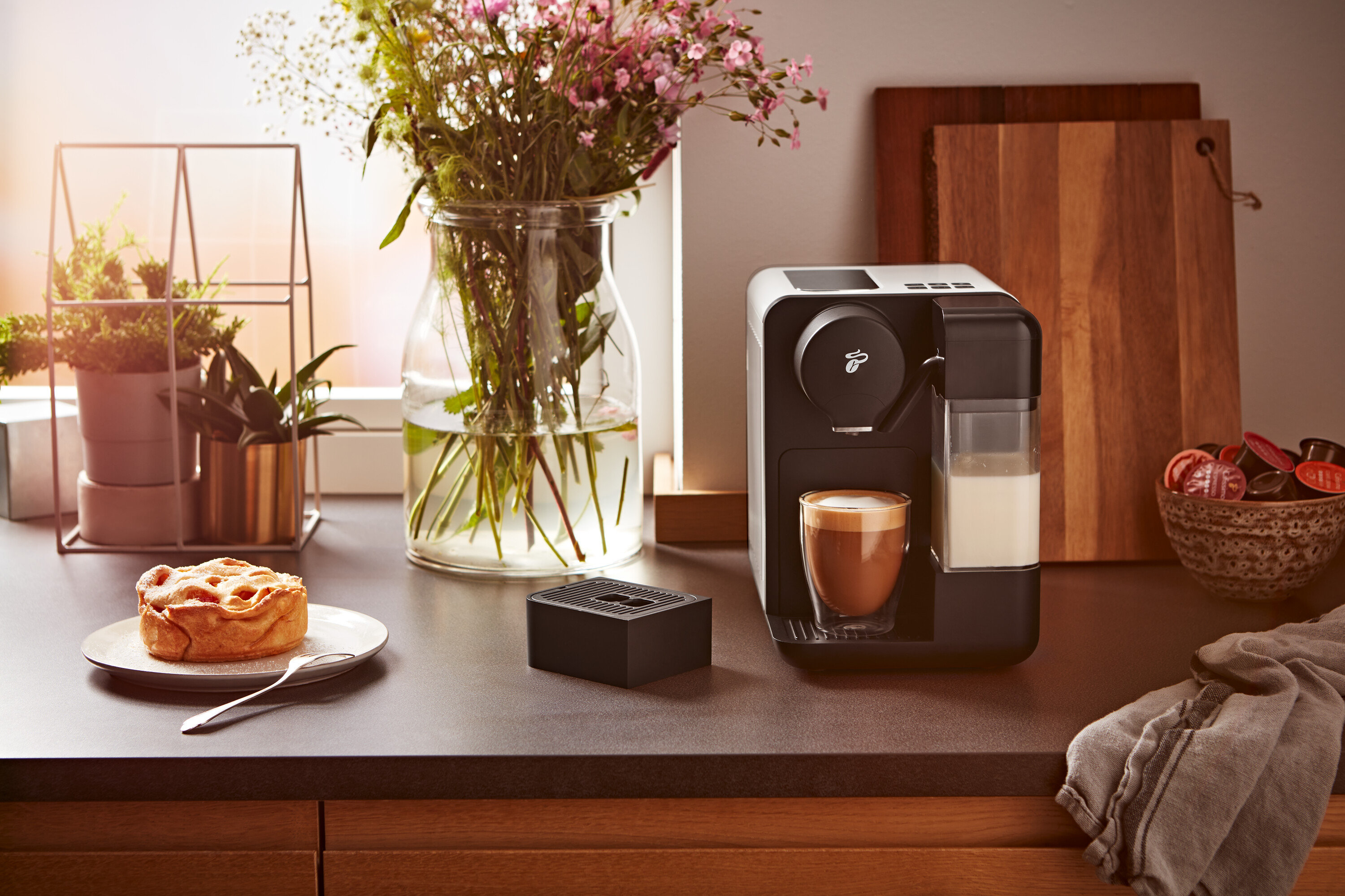 Na linke stojí kávovar vedľa pohára mlieka a šálky kávy. V pozadí váza s kvetmi a dosky na krájanie.