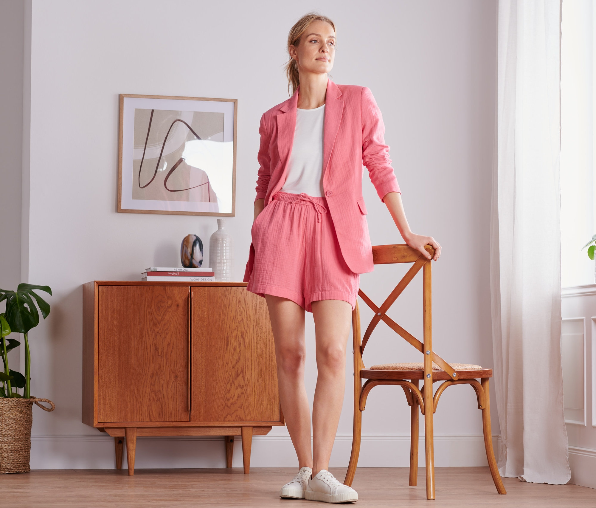 Žena pózuje v koralovom mušelínovom saku a šortkách. Opiera sa o stoličku vedľa okna.