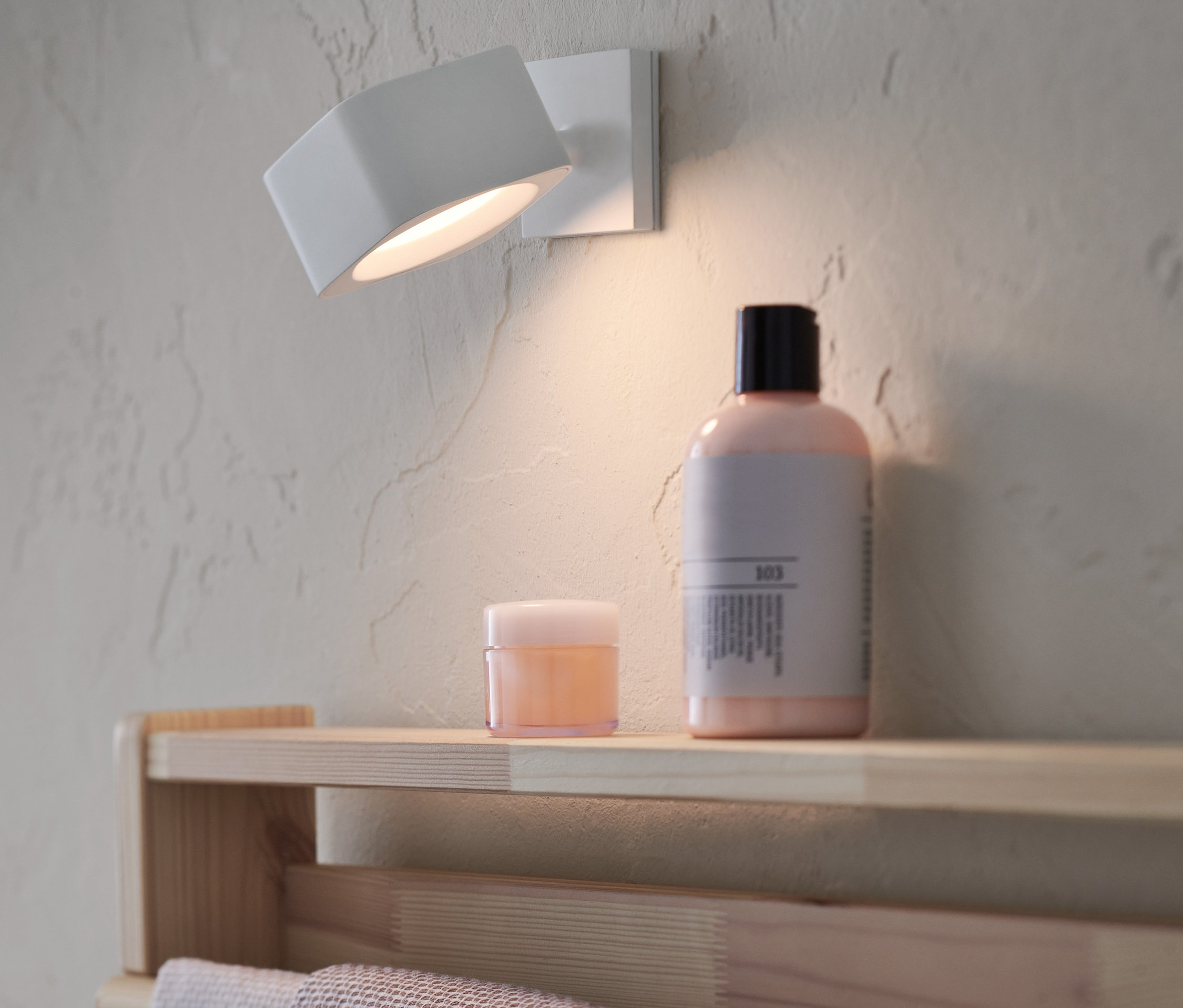 Na stene je pripevnené nástenné LED svietidlo na batérie. Na poličke pod ním stojí kozmetika.