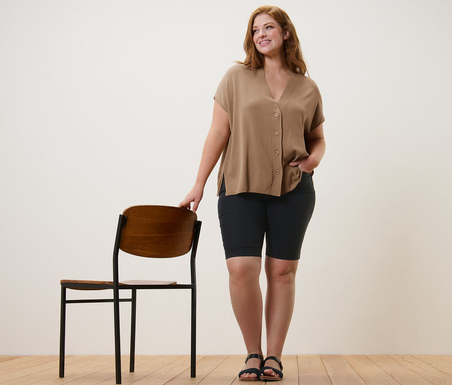 Žena stojí vedľa stoličky a má na sebe béžový kardigán s krátkymi rukávmi a čierne bengalínové bermudy.