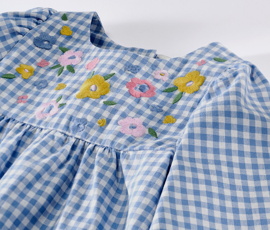 Detail detských tkaných šiat s modro-bielym károvaným vzorom a výšivkou kvetov.