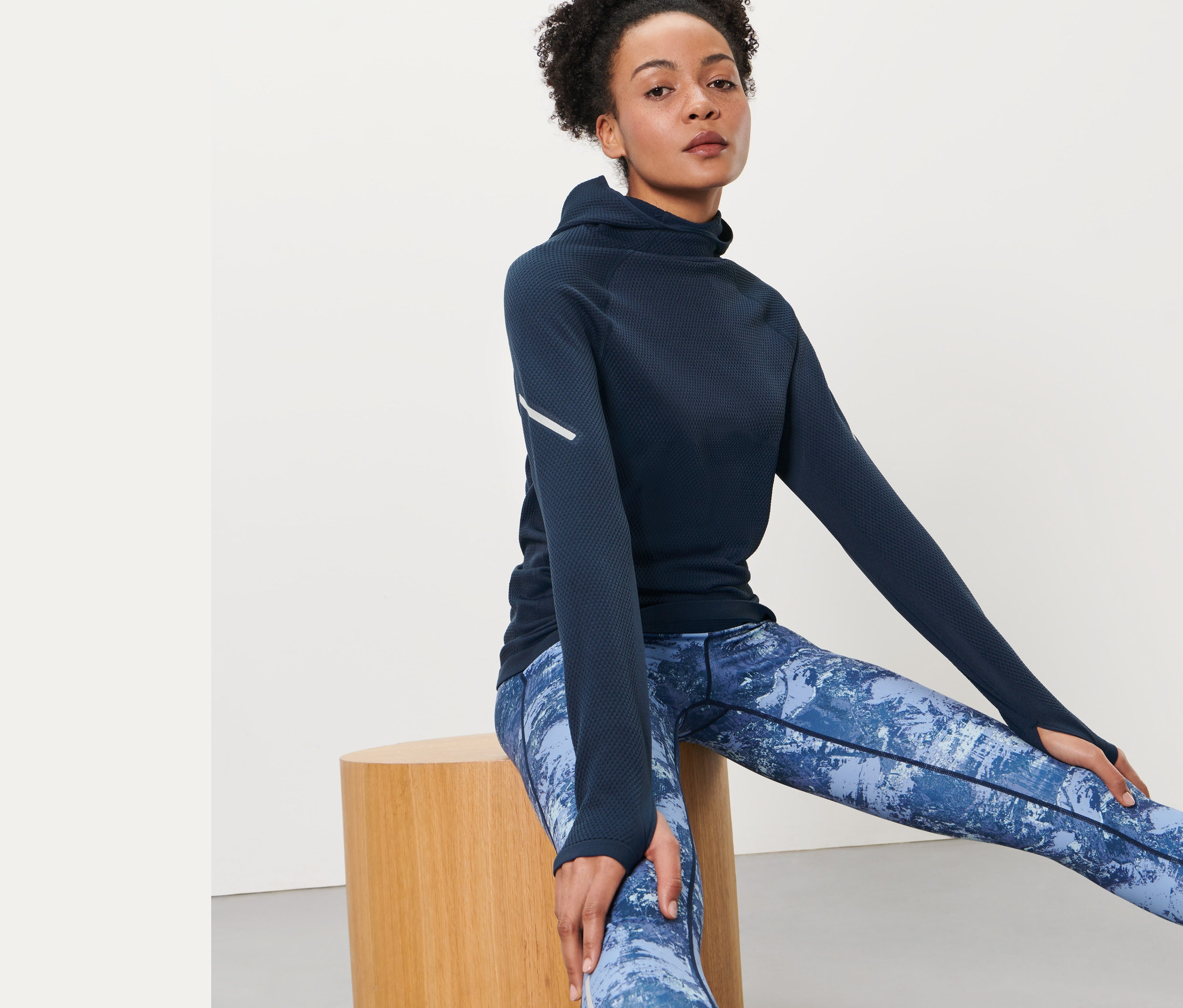 Žena sedí na drevenej stoličke a má na sebe bezšvové funkčné bežecké tričko s kapucňou a modré vzorované legíny.