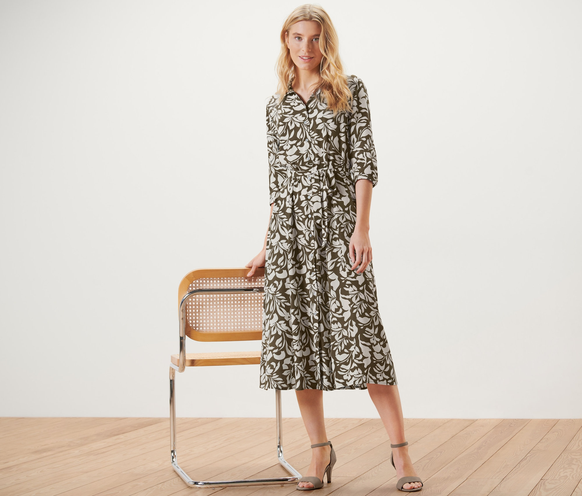 Blondína pózuje vedľa stoličky a má na sebe košeľové šaty s potlačou.