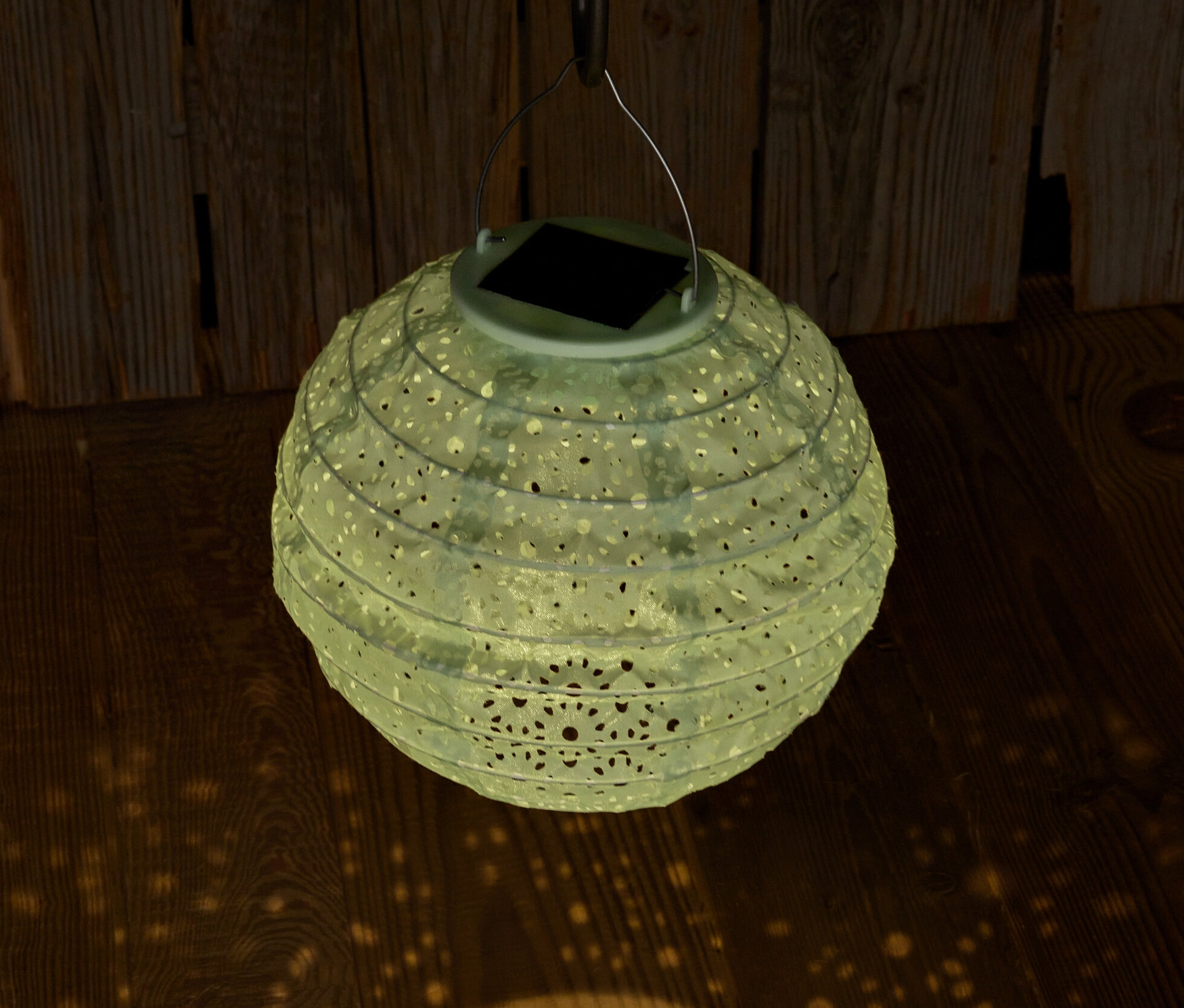 Zavesený solárny lampión s ozdobnými otvormi premieta svetlo na drevenú podlahu.