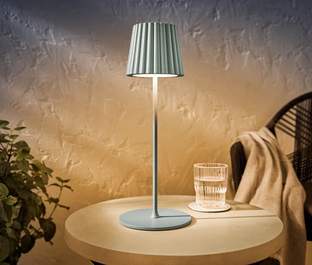 Šalviovo sfarbená LED akumulátorová stolová lampa do exteriéru stojí na okrúhlom stolíku vedľa pohára vody.