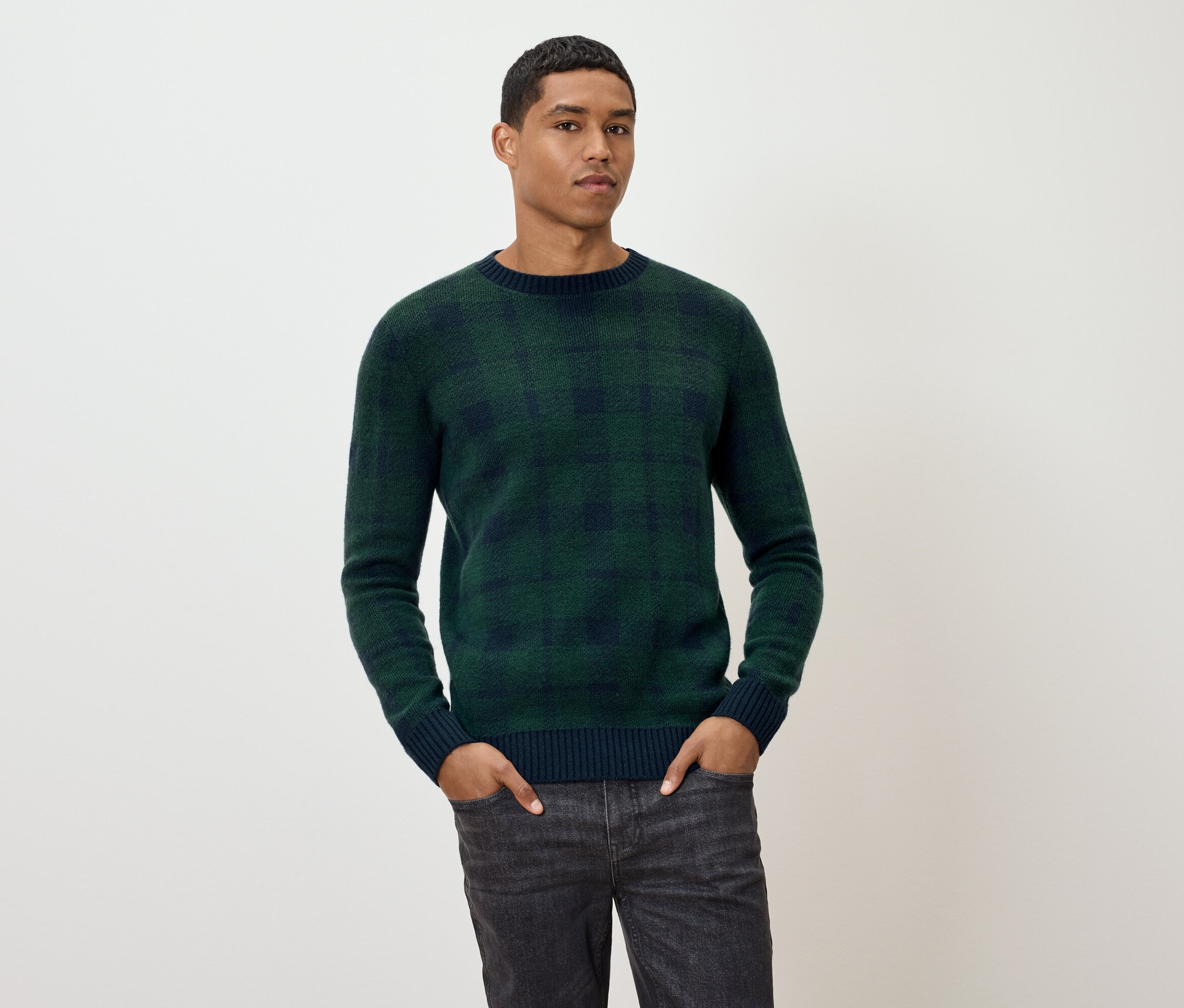 Muž pózuje v zelenom kockovanom svetri a sivých džínsoch.
