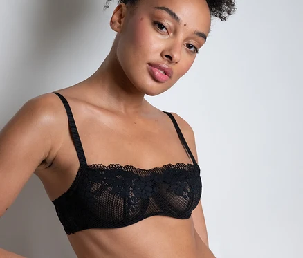 Žena s vyčesanými kučeravými vlasmi má na sebe čiernu čipkovanú bandeau podprsenku Passionata »Nina« a zhodné nohavičky, s rukou na boku.