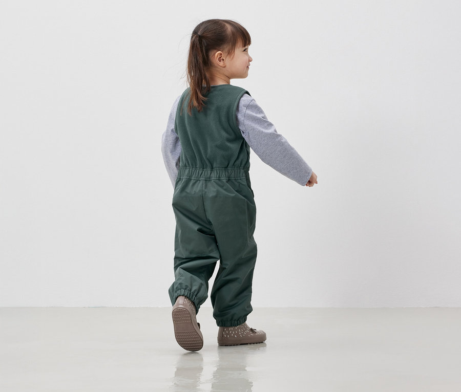 Dievča v zelenom overale a sivom tričku kráča po bielej podlahe.