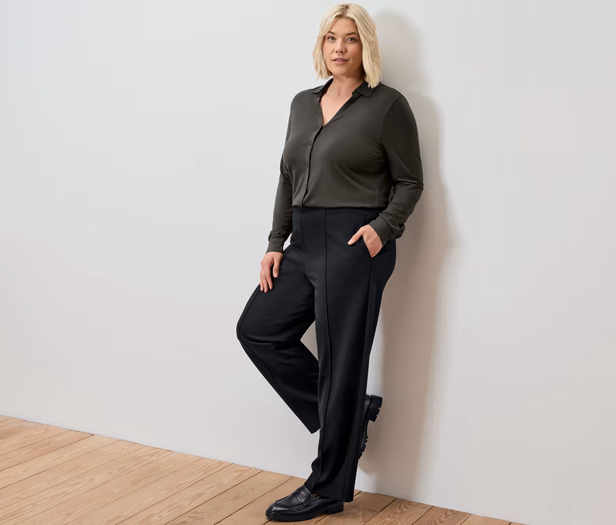 Blond žena opretá o stenu má na sebe čierne džersejové nohavice so zámikom a blúzkové tričko s gombíkovou légou.