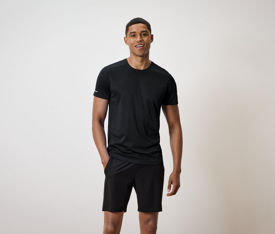 Usmievajúci sa muž stojí a má ruky vo vreckách čiernych šortiek a má na sebe čierne tričko.