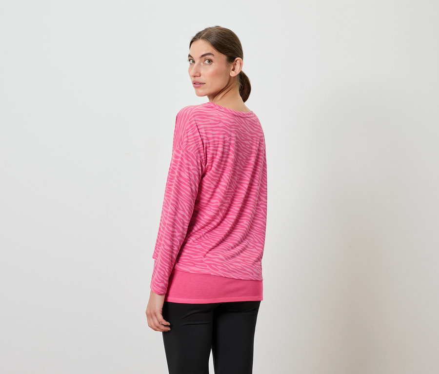 Žena sa pozerá cez plece, má na sebe ružový top s dlhým rukávom a čierne nohavice.