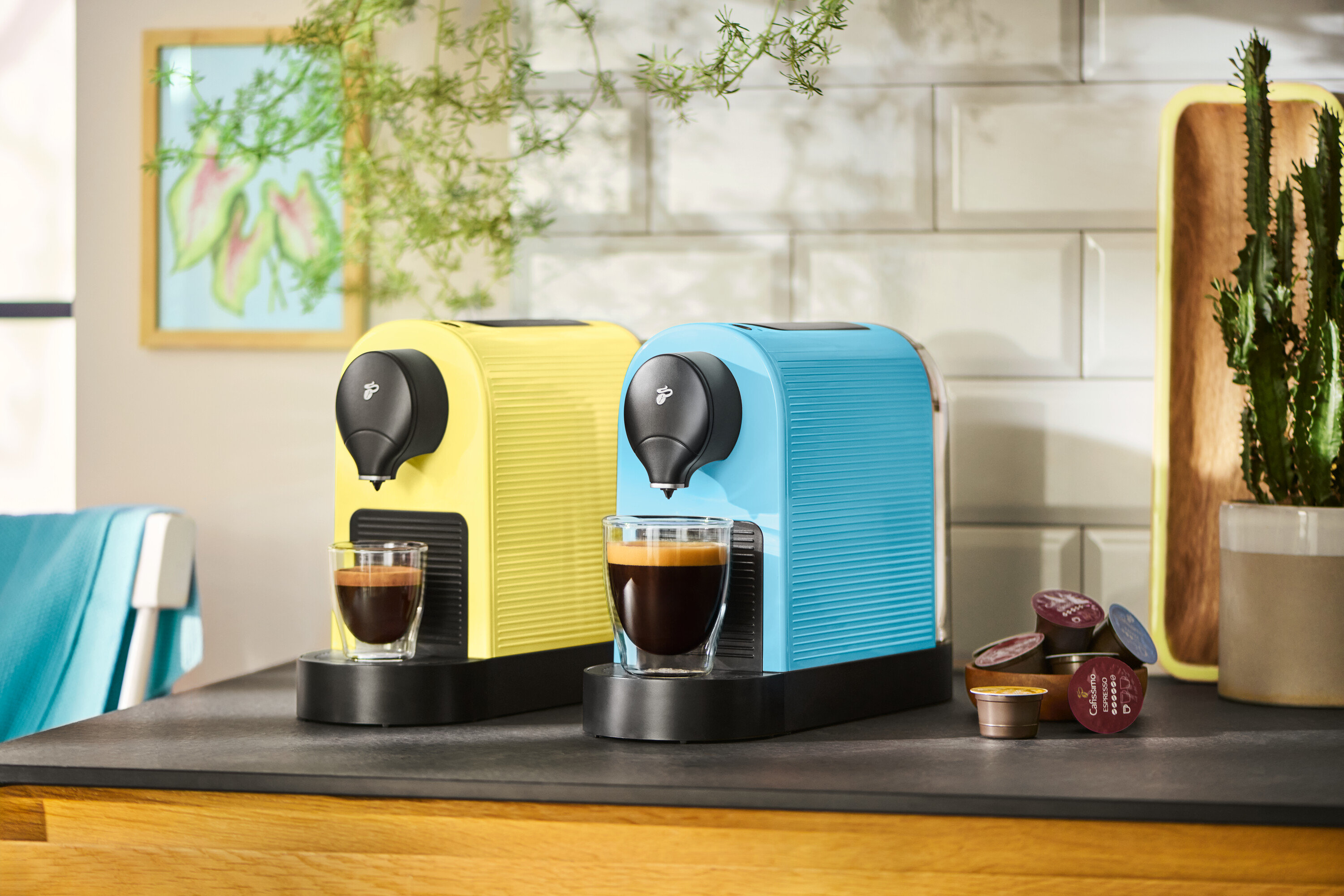 Dva kávovary v žltej a modrej farbe stoja na kuchynskej linke s dvoma šálkami kávy a kávovými kapsulami.