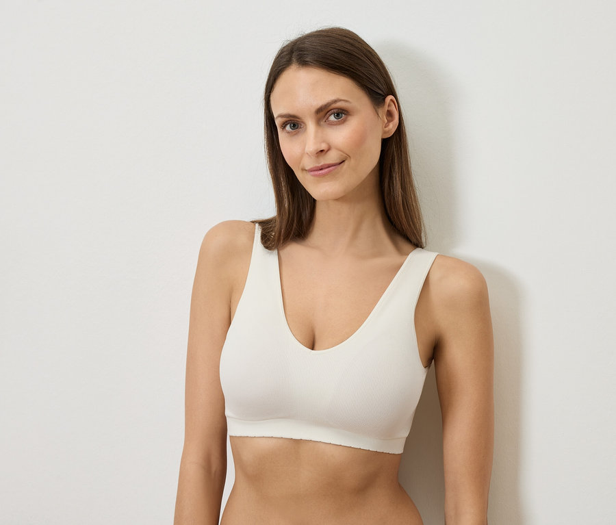 Žena s dlhými hnedými vlasmi v bielej podprsenke sa opiera o bielu stenu.