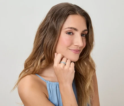 Žena s vlnitými vlasmi a v modrom tope sa dotýka prsta s prsteňom na svetlom pozadí.