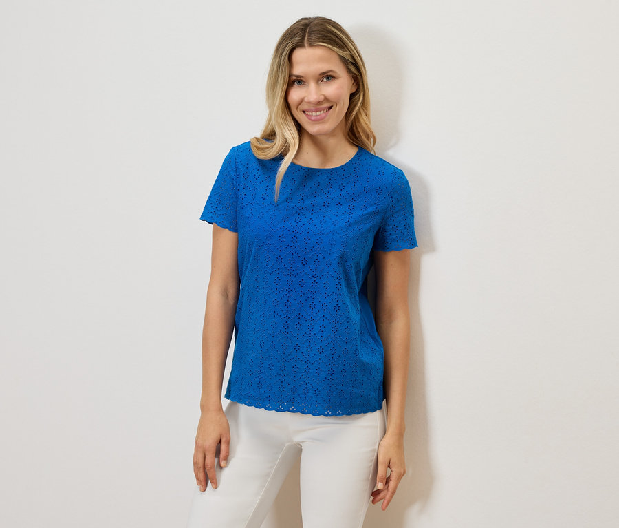 Žena sa usmieva a má na sebe modré tričko s krátkym rukávom a dierkovaným vzorom a biele nohavice na bielom pozadí.