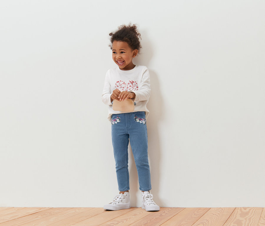 Usmievavé kučeravé dievča s béžovou kabelkou, blúzkou s dlhým rukávom a modrými džínsami s kvetinovou výšivkou, stojace pred bielou stenou.