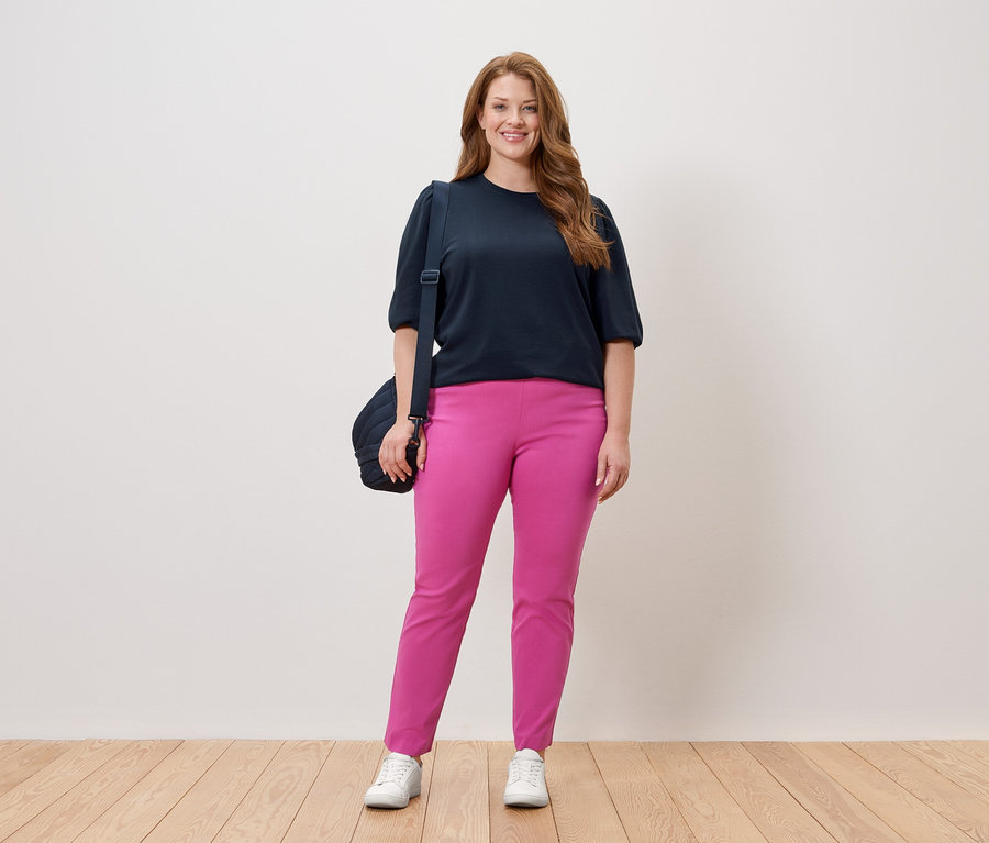 Žena stojí v tmavomodrom tope a ružových nohaviciach s taškou cez rameno pred bielou stenou.