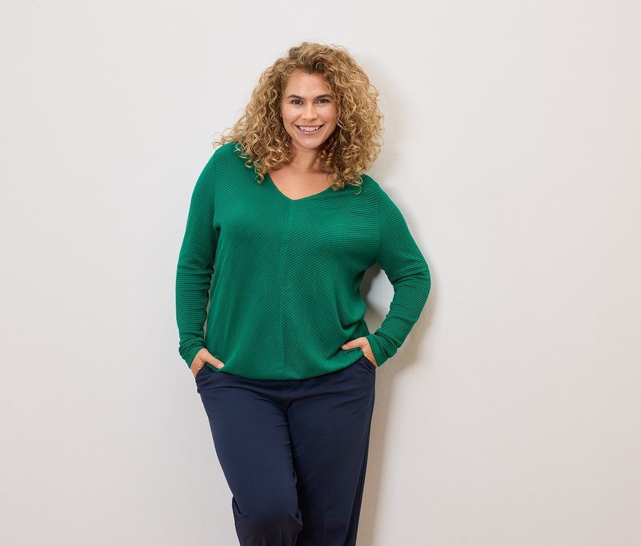 Žena s kučeravými vlasmi má na sebe zelený sveter a tmavomodré nohavice, ruky vo vreckách.