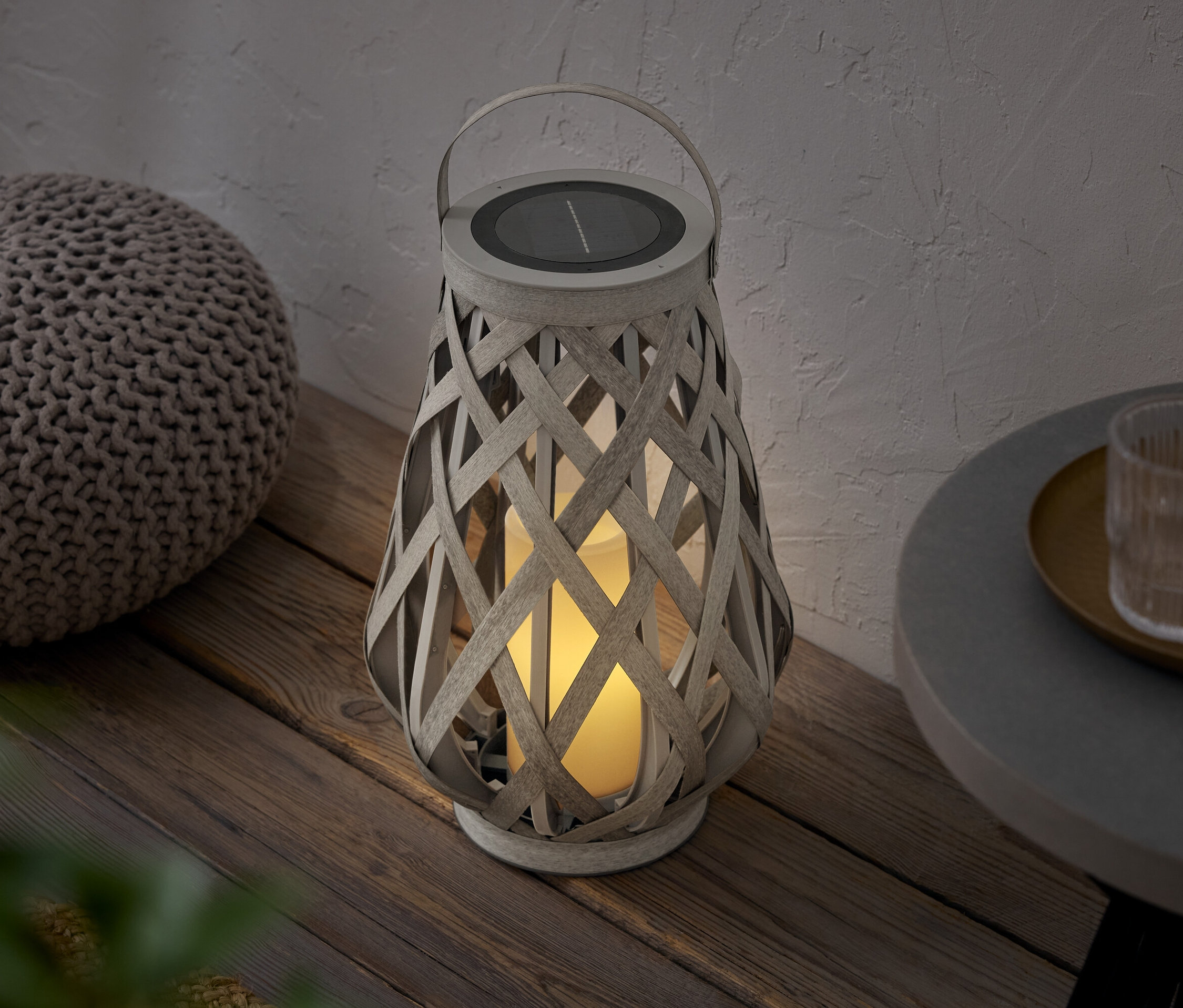 Pletená lampa so solárnym panelom stojí vedľa pufu na drevenej podlahe.